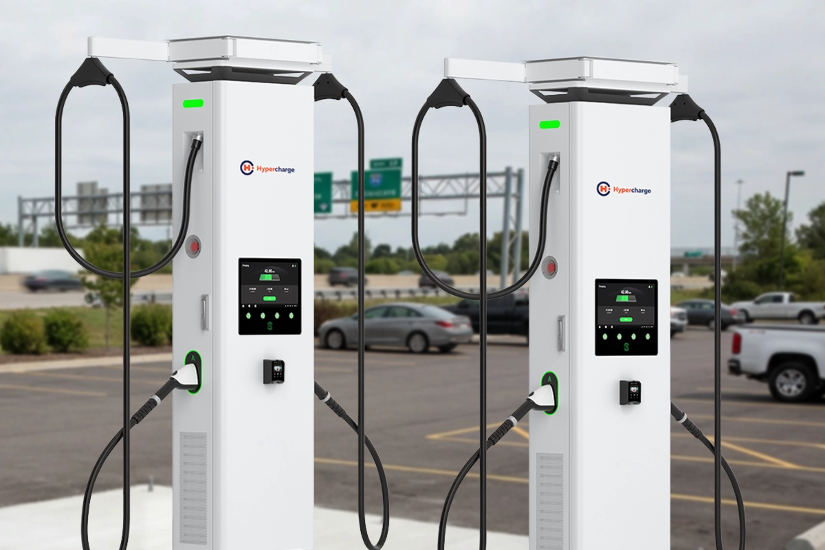 Two Hypercharge white electric vehicle charging stations with digital screens and charging cables stand in a parking lot near a highway. Two Hypercharge white electric vehicle charging stations with digital screens and charging cables stand in a parking lot near a highway.