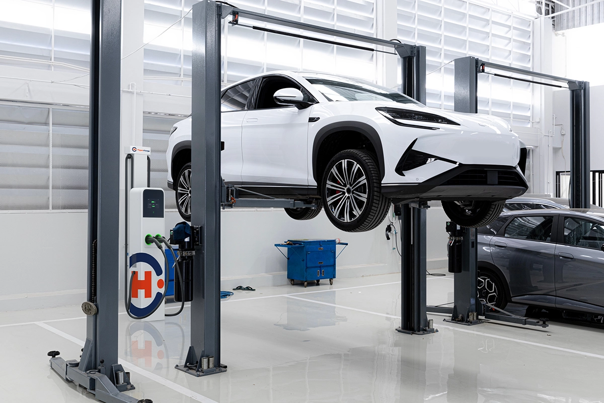 A white electric car is elevated on a hydraulic lift inside a bright, modern automotive service garage, with tools and equipment visible in the background—prompting the question: How Much Does It Cost to Maintain an EV?.
