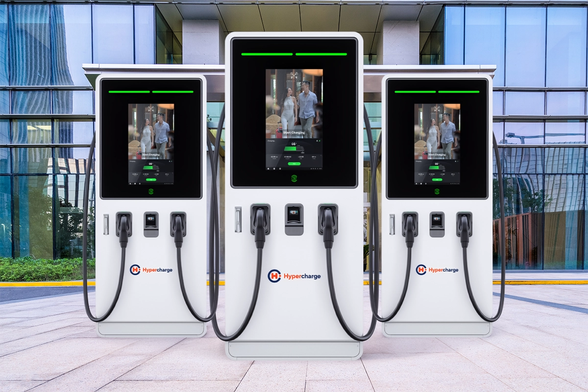 Three Hypercharge EV charging stations stand outdoors in front of a modern glass building, each screen displaying a person walking inside a hotel lobby—showing why leading hotels need EV charging stations for added guest convenience.