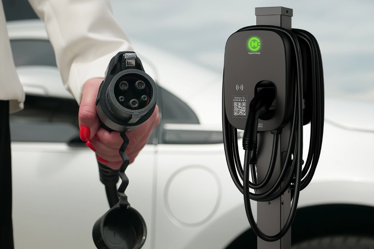 A person with red nail polish holds an electric vehicle charging plug near a Hypercharge station, featuring a green icon and coiled cable, as a white car waits.
