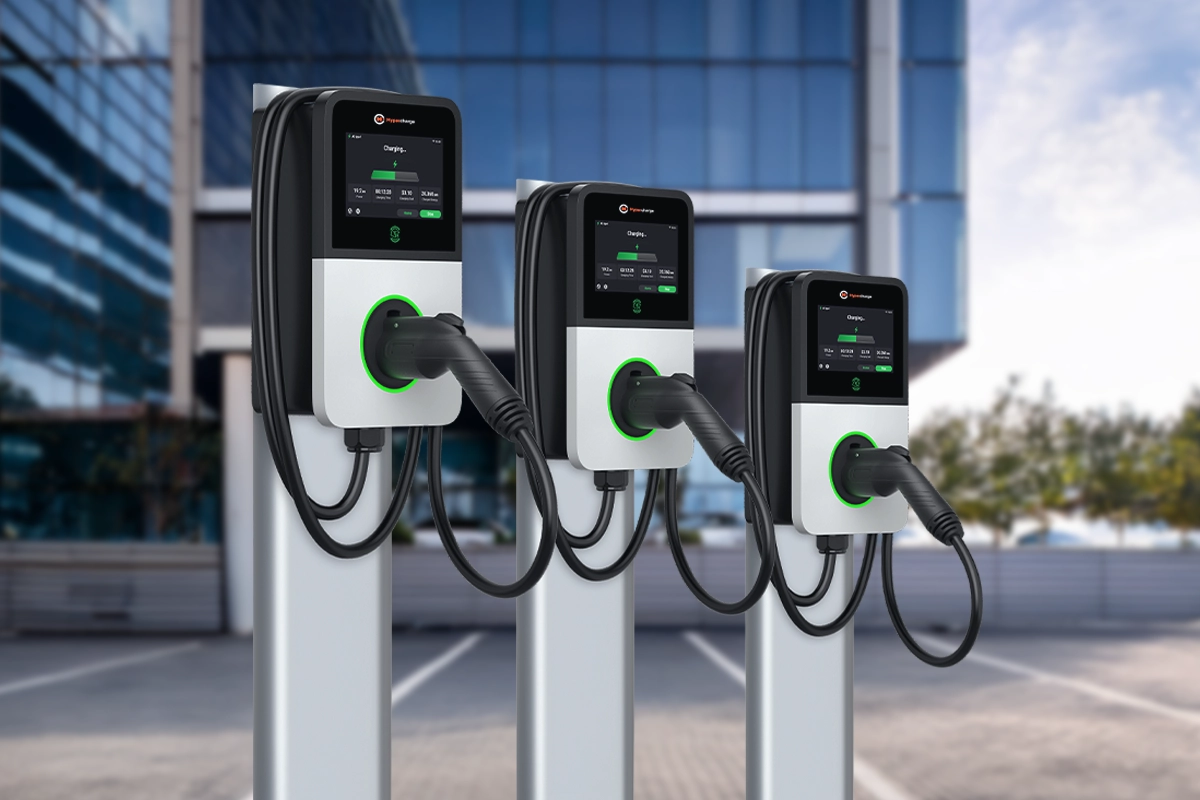 Three electric vehicle charging stations with digital displays are installed outdoors in a parking lot near a modern glass building, meeting the rising demand for workplace EV charging amid a backdrop of trees. Three Autel MaxiCharger AC Pro: electric vehicle charging stations with digital displays are installed outdoors in a parking lot near a modern glass building, meeting the rising demand for workplace EV charging amid a backdrop of trees.