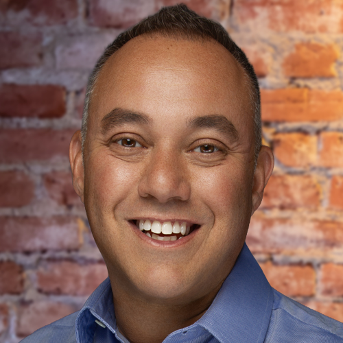 Malcolm Davidson, CFO & CPA smiling wearing a blue collared shirt stands in front of a brick wall background.