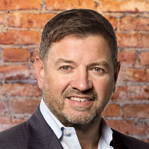 Tony Geheran, former EVP & Chief Operations Officer at TELUS, photographed in a dark suit in front of a brick wall. A man with short brown hair, trimmed beard, and light eyes is smiling. He is wearing a dark suit jacket over a light blue shirt, standing in front of a brick wall background.